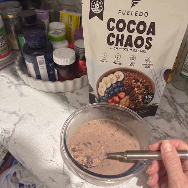 A hand holding a spoon with prepared oatmeal in front of a stand-up pouch of Fueledo Cocoa Chaos high protein oat mix on a kitchen counter.