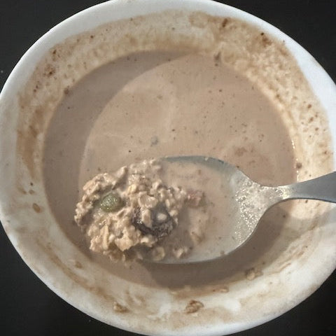 A white bowl containing Fueledo High Protein Oatmeal, with a spoon holding a scoop of the same mixture above it, set on a dark surface.