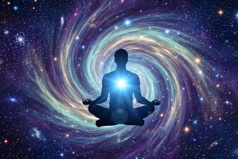 A digital illustration of a dark human silhouette sitting in a meditative lotus pose, floating in deep space. A bright, glowing white light radiates from the figure's chest, casting a soft blue aura around the upper body. The background is a vibrant, swirling spiral galaxy filled with shades of blue, teal, purple, and gold cosmic dust, surrounded by a dense field of twinkling stars and distant celestial bodies.