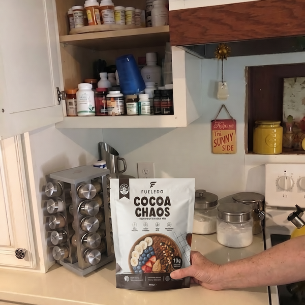 A person's hand holds a bag of Fueledo Cocoa Chaos in a kitchen setting with white cabinets, spice jars, and a stove in the background.